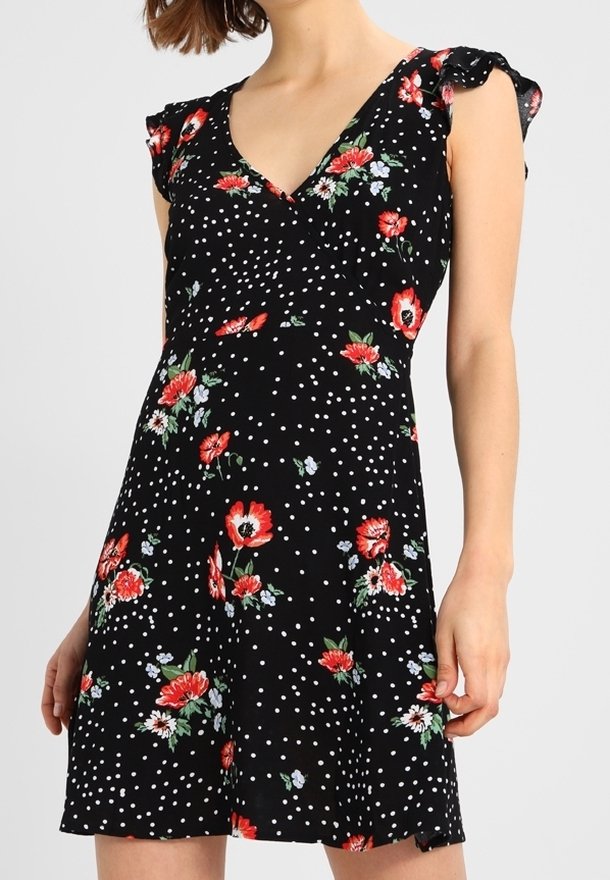 Femme portant une robe noire à pois blancs et motifs floraux rouges, avec un col en V et des manches courtes.