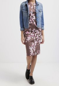 En denimjakke over en tætsiddende brun blomsterkjole med lyserøde blomster. Sorte sko, glat tekstur og knælang silhuet.