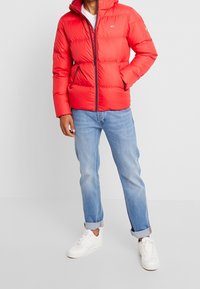 Rød puffjakke med hætte, med lynlås foran og to sidelommer. Båret over en hvid skjorte og parret med lyseblå jeans og hvide sneakers.