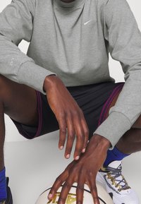 Camiseta gris de manga larga de Nike, pantalones cortos negros con acentos morados. Una pelota de baloncesto está en el suelo, con las manos apoyadas en ella. Se pueden ver zapatillas deportivas.