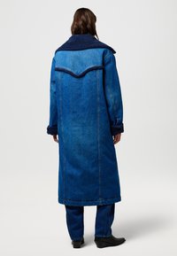 Długi płaszcz dżinsowy z granatowym kołnierzem z ekologicznego kożucha. Charakteryzuje się prostym krojem, bocznymi kieszeniami oraz niebieską teksturą dżinsu z detalami przeszyć.