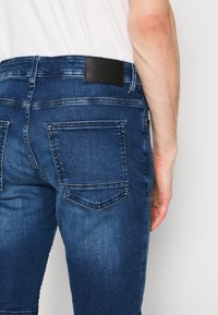 Jeans in denim blu con una texture liscia, un classico design a cinque tasche e una patch in cuoio nero in vita con il logo del marchio.