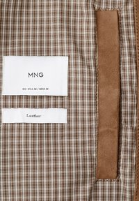 Braun-beiges karriertes Stoffmuster mit zwei weißen Etiketten, die "MNG EU-USA M / MEX M" und "Leder" zeigen, neben einem braunen Lederstreifen.