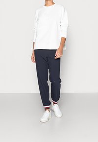 Sudadera blanca con logotipo en relieve, pantalones tapered marinos con puños acanalados en rojo y blanco, y zapatillas blancas con suelas texturizadas.