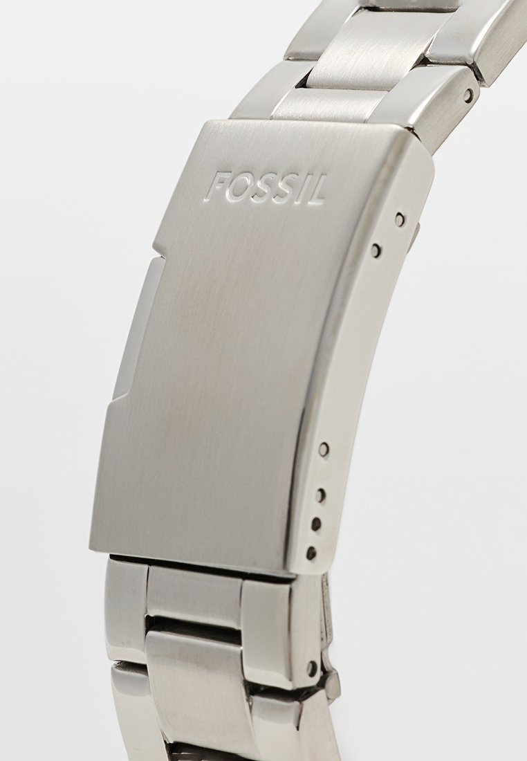 Reloj de acero inoxidable plateado con un acabado pulido y cepillado. Presenta un logotipo de Fossil y eslabones ajustables con agujeros visibles para el tamaño.