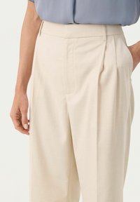Pleated beige trousers made of soft fabric, featuring side pockets and a tailored fit, shown with a blue top and hand positioned on the hip.