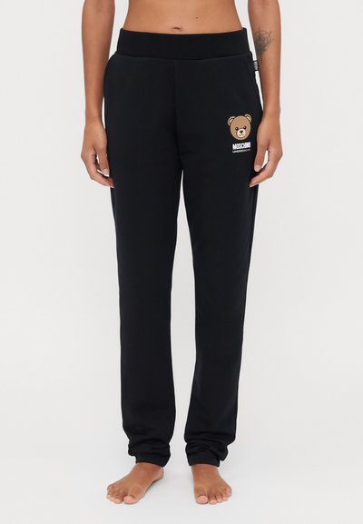 Pantalon de survêtement noir avec une taille côtelée, présentant un graphic d'ours et le texte "Moschino Underbear" sur la jambe gauche. Tissu doux.