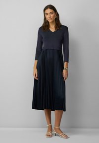 Marineblaues plissiertes Kleid mit taillierter Oberweite und V-Ausschnitt, kombiniert mit weißen, geschnallten Absätzen. Strukturierter Strickstoff oben, fließender Rock darunter.