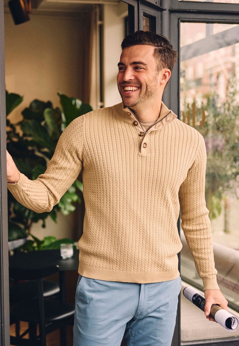 Beige gebreide trui met een gestructureerd patroon, voorzien van een halve knoopsluiting en lange mouwen, gecombineerd met lichtblauwe broek en een opgerolde tijdschrift in de hand.