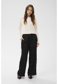 Kremowy kardigan z długim rękawem zapinany z przodu na guziki, zestawiony z czarnymi teksturowanymi spodniami z frędzlami, posiadającymi ukośny wzór i boczne kieszenie.