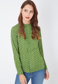Blusa verde de manga larga con cuello, con un patrón de lunares blancos. Panel superpuesto en la parte delantera y textura de tela suave.