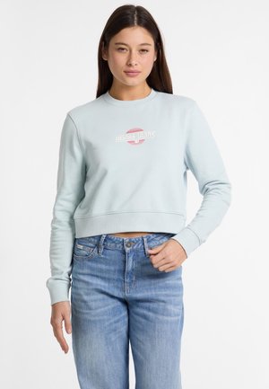 Lichtblauwe cropped sweatshirt met lange mouwen, ribgebreide manchetten en een geborduurd "Guess Jeans" logo in rood en wit. Draag met blauwe jeans.