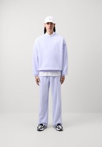 Gaiši violets pārmērīgs sporta krekls ar izšūtu tekstu, apvienots ar atbilstošām gaiši violetām biksēm. Baltā cepure un melnbaltas rakstainas kurpes.