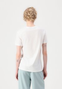 White cotton t-shirt with short sleeves, round neckline, and a plain design, paired with light blue trousers, viewed from the back.