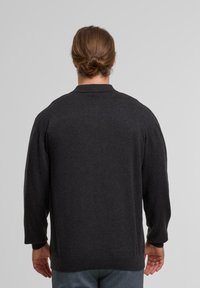 Person mit zurückgebundenem Haar, der einen dunkelgrauen langärmeligen Pullover und hellgraue Hose trägt und mit dem Rücken zu einem einfarbigen Hintergrund steht.