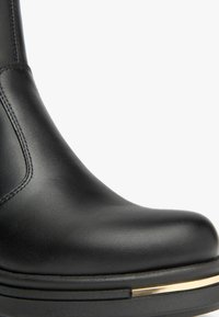 Botte de cheville en cuir noir avec un bout rond, un talon surélevé et un accent en or le long de la semelle. Texture lisse et détails de couture soignés.