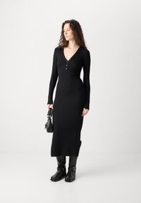Black ribbed knit dress with long sleeves, V-neckline, and button detail. Paired with black boots and a small black handbag.