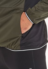 Giacca sportiva di un verde scuro e nero con tasca con zip, caratterizzata da bande bianche lungo l'orlo e i polsini, realizzata in un tessuto leggero e liscio.