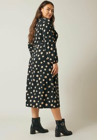 Schwarzkleid mit großen goldenen Punkten, langen Ärmeln und tailliertem Bund. Kombiniert mit schwarzen Stiefeletten mit einem klobigen Absatz und Schnallen.