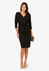 Schwarz, knielanges Kleid mit V-Ausschnitt, Dreiviertelärmeln, tailliertem Detail und glattem Stoff. Kombiniert mit einer gestreiften Clutch und nudefarbenen High Heels.