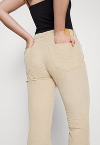 Beige manchesterbyxor med en strukturerad yta, med bakfickor och en gul tagg vid midjan, visade från en sidovinkel.