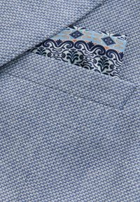 Modrý štruktúrovaný materiál saka so vzorom cikcak, doplnený kontrastnou vreckovkou v modrej, bielej a oranžovej kvetinovej potlači.