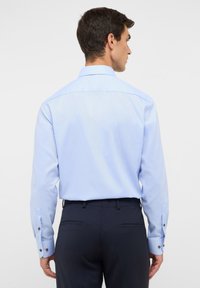 Chemise bleu clair avec un col classique, des manches longues et des poignets boutonnés. La chemise présente une texture lisse et une coupe ajustée.