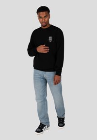 Sudadera negra con un gráfico de una figura de dibujo animado en el lado izquierdo del pecho, combinada con jeans de azul claro y zapatillas negras con acentos blancos.