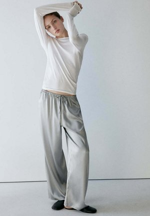Frau, die die Arme über den Kopf streckt, trägt ein weißes Langarmshirt, lockere silberne Satin-Hosen und schwarze Slip-On-Schuhe vor einem schlichten hellen Hintergrund.