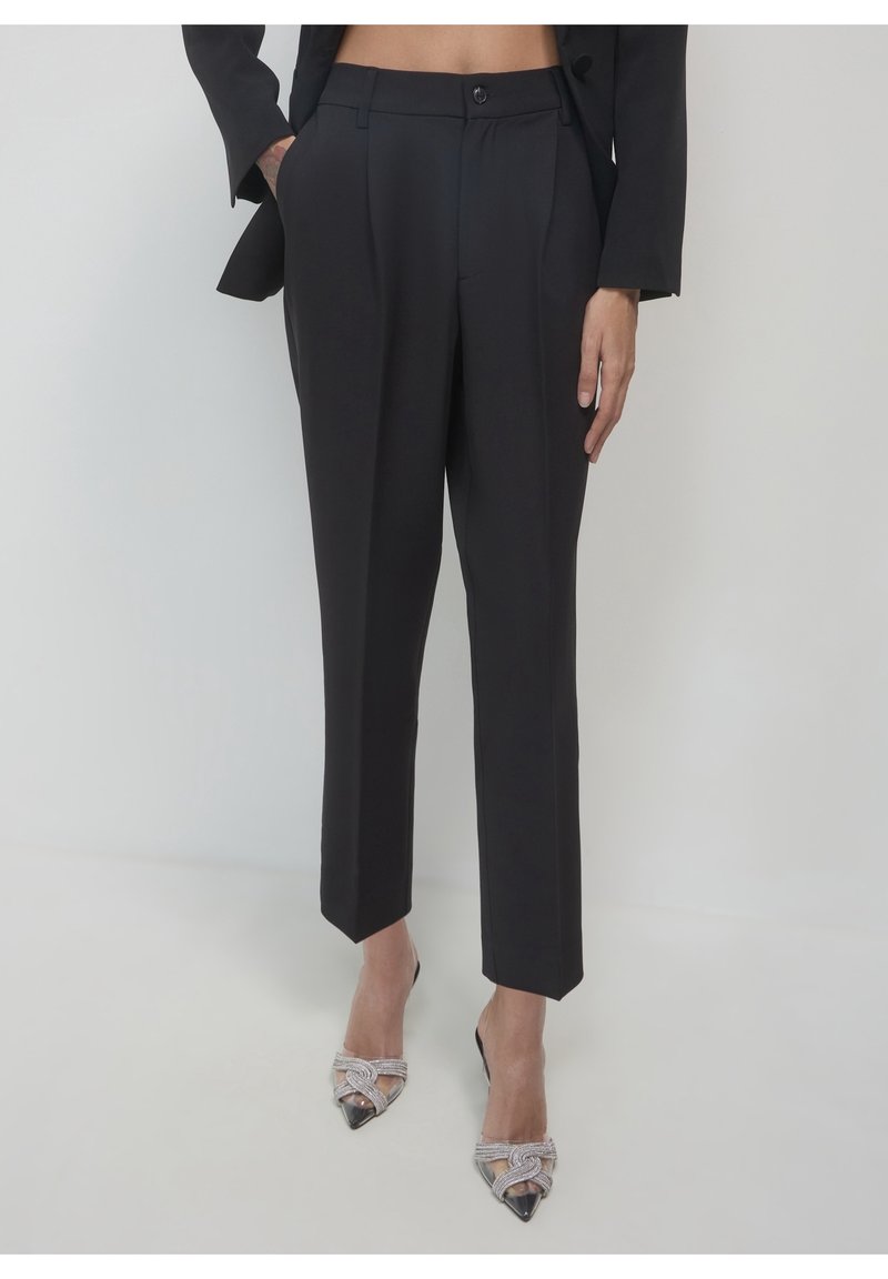 Femme portant un pantalon noir taille haute ajusté et un blazer noir raccourci à la taille, associé à des escarpins pointus ornés.