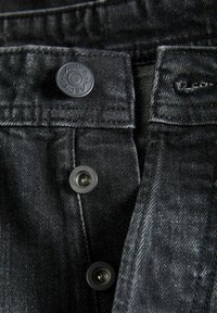 Tela de mezclilla negra con un primer plano de broches metálicos y un detalle de botón de marca en la cintura. La textura está ligeramente desgastada.