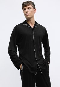 Camisa de pijama de manga larga negra con cuello y ribete blanco, con botones en el frente y un logotipo sutil en el pecho.