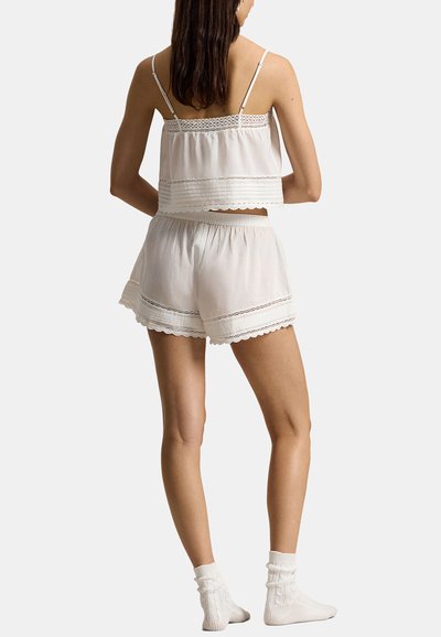 Woman wearing white lace-trimmed camisole and shorts set with white ankle socks, standing with back facing forward against plain background.