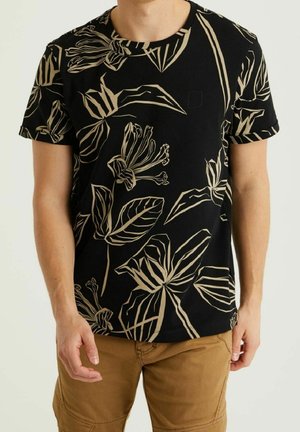 Mand iført sort T-shirt med beige tropiske blade- og blomsterprint og brune bukser, stående mod en ensfarvet baggrund.