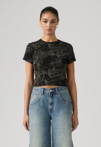 Κοντοκομμένο μαύρο t-shirt με υφή εκτύπωσης που παρουσιάζει διάφορα λογότυπα· συνδυασμένο με φαρδιά μπλε τζιν παντελόνια. Κοντά μανίκια και στρογγυλός λαιμός.