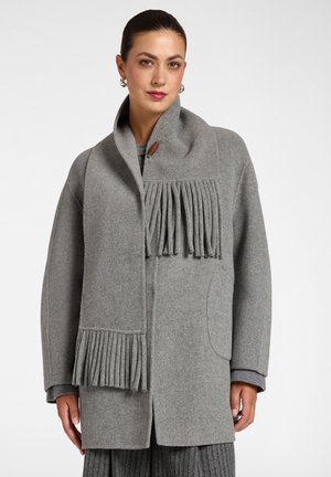 Donna che indossa un cappotto di lana grigio con una sciarpa a frange abbinata drappeggiata sulle spalle su uno sfondo semplice.