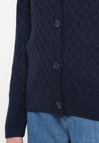 Cardigã malha marinha com padrão em diamante, com punhos e bainha canelados, apresenta botões escuros na frente, feito de lã macia e texturizada.