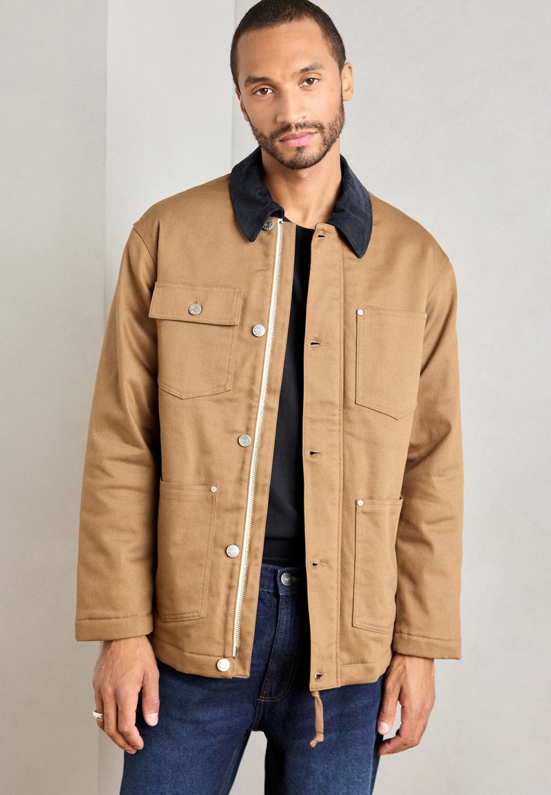 Hombre vistiendo una chaqueta beige con un collar negro, cierres de botones y cremallera, dos bolsillos en el pecho y jeans azules, de pie contra una pared lisa.