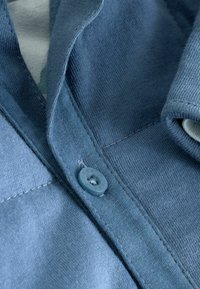 Zbliżenie na niebieską dzianinę z pojedynczym guzikiem i widocznym szyciem, ukazujące część złożonego rękawa oraz teksturowany materiał.