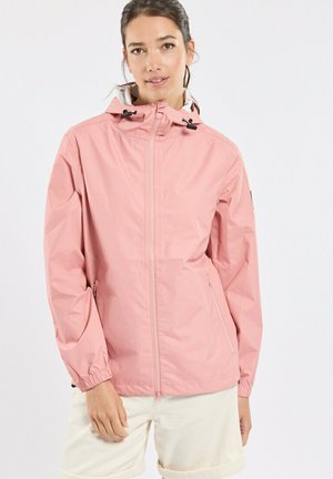 Femme portant une veste à capuche rose clair avec fermeture éclair avant et poches, associée à un short de couleur crème.