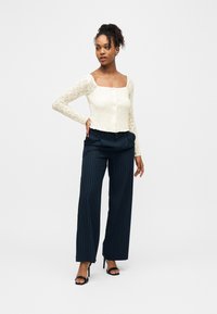 Γυναίκα όρθια με το χέρι στη μέση, φοράει κρεμ τοπ με τετραγωνισμένη λαιμόκοψη από δαντέλα, παντελόνια σε σκούρο μπλε με λεπτές ρίγες και μαύρα σανδάλια με τακούνι.