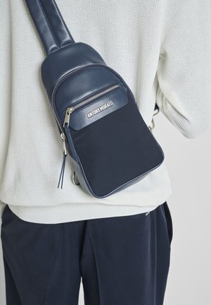 Sac bandoulière bleu marine avec fermeture éclair, porté sur un pull blanc texturé et un pantalon foncé, affichant le nom de la marque "Antony Morato" à l'avant.