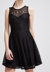 Schwarzes Spitzenkleid mit einem taillierten Oberteil, einem gewellten Ausschnitt und einem ausgestellten Rock, filigranen Blumenmustern und transparenten Details an der oberen Hälfte.