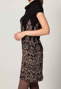 Knielange jurk met korte mouwen en een col hals, met een zwart en lichtbruin damastpatroon op een donkere achtergrond. Zachte stoftextuur.