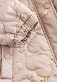 Quiltet jakke i blød beige stof med blomstrede detaljer, samlede ærmedesign og runde knapper til lukning.