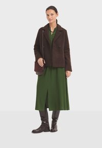 Veste croisée en laine brun mélangé sur une robe longue verte avec une fente sur le côté, associée à des bottes en cuir brun mi-mollet. Sac à épaule brun.