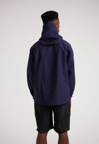 Marineblå vandtæt jakke med hætte, der har en glat tekstur og justerbare manchetter, iført over sorte cargoshorts med sidelommer.