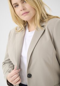 Blazer beige chiaro con un design strutturato, revers a scendere e un singolo bottone nero. Indossato sopra una t-shirt bianca, che mostra una texture del tessuto liscia.