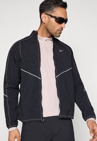 Črno lahko jakno s belim poudarkom šivov, z zadrgo in stranskimi žepi, nošeno preko svetlo roza majice z visokim ovratnikom.