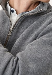 Il felpa grigia con zip presenta una texture morbida, polsini a coste e un colletto alto. Sotto, una camicia con colletto aggiunge un motivo a contrasto.